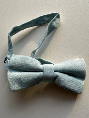 Ljusblå fluga i bomulls-denim- helt NY - Snygg ljusblå fluga i mjukt bomullstyg med klassisk form och justerbart band. Perfekt för att ge din outfit en stilren touch. Enkel att knäppa och passar till många olika looks.