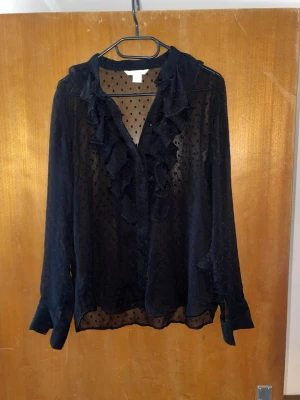 Svart transparent blus med volanger H&M - Snygg svart transparent blus från H&M med volangdetaljer längs knäppningen och små prickar i tyget. Blusen har lång ärm och är tillverkad i ett lätt och luftigt material som ger en cool look.