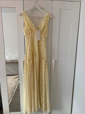 Gul långklänning från Adoore  - Ljusgul långklänning från Adoore  med v-ringning och smala axelband. Klänningen har ett subtilt blommigt mönster och är tillverkad i ett lätt, luftigt material som passar perfekt för varma dagar. Modellen är feminin med markerad midja och voluminös kjol.