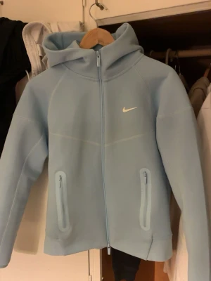 Nike nocta tech fleece baby blue  - Snygg ljusblå hoodie från Nike med huva och dragkedjeförsedd ficka på sidan. Hoodien har diskreta vita loggor och broderade detaljer, samt mjukt material som känns skönt mot huden. Perfekt för en avslappnad och sportig stil. Passformen är som S men funkar både för folk med storlek S och M.