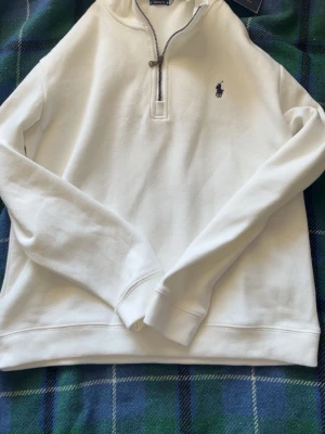 Vit half-zip hoodie från Ralph Lauren - Snygg vit hoodie från Ralph Lauren med half-zip dragkedja och klassisk broderad logga på bröstet. Tillverkad i mjukt bomullsmaterial och har långa ärmar samt ribbade muddar. Perfekt för en clean och stilren look.