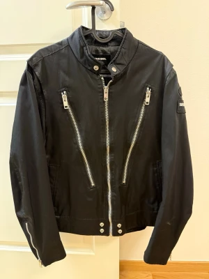 Svart bikerjacka från Diesel - Svart skinnjacka från Diesel med coola silverfärgade dragkedjor, ståkrage och två snedställda bröstfickor. Jackan har detaljerade knappar vid nederkanten och patch på ärmen för extra attityd. Perfekt för dig som vill ha en edgy look.
