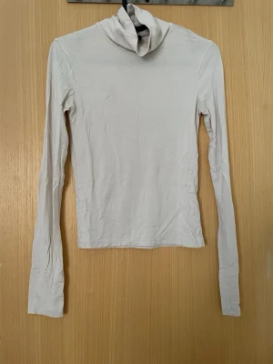 Vit topp med lång ärm - Säljer en enkel och stilren vit topp  med lång ärm. Toppen har en hög krage och är tillverkad i ett mjukt material som sitter tight mot kroppen. Perfekt att matcha med jeans eller kjol för en clean look.