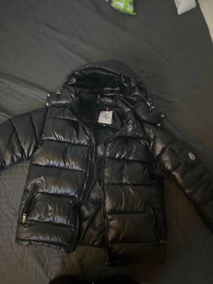 Svart pufferjacka från Moncler - Fick den i födelsedagspresent, Snygg svart pufferjacka från Moncler med glansig finish och vadderad design. Jackan har huva, dragkedja framtill och två fickor med dragkedja. Klassisk Moncler-logga på ärmen och detaljer i silver. Perfekt för kalla dagar och riktigt trendig look.