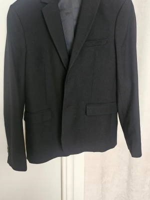 Svart klassisk kavaj med fickor - En svart kavaj med två knappar framtill, klassiska slag och tre fickor med lock. Kavajen har en stilren och tidlös design med slät yta och helfodrad insida. Perfekt för en dressad look och passar till många olika outfits.