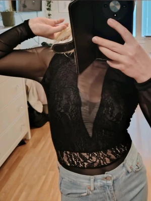 Svart mesh och spets body med v-ringning🖤  - Snygg svart långärmad body med transparenta meshärmar och spetsdetaljer😍 Djup v-ringning framtill och figurnära passform😌 Perfekt att styla med jeans eller kjol för en trendig look! Aldrigt använt! 