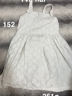 Vit blommig sommarklänning - Söt vit klänning med broderade blommor och axelband med knutdetalj. Klänningen har en lätt utställd kjol och är perfekt för varma dagar.🌸Diffusa fläckar och två hål. Storlek 152