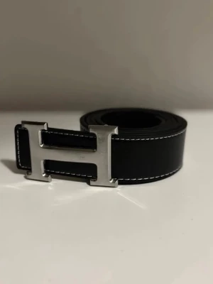 Svart skärp med H-spänne i silver - Stilrent svart skärp från  med ett ikoniskt H-format spänne i silverfärgad metall. Skärpet är gjort i skinn med vit kontrastsöm längs kanterna och har en klassisk, tidlös design som passar till många outfits.