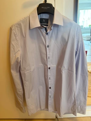 Ljusblå skjorta från Savvy Citizen  - Snygg ljusblå skjorta från Selected Homme med klassisk krage och svarta knappar. Skjortan har lång ärm och är tillverkad i bomull, perfekt för en stilren look.