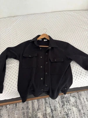 Svart overshirt från Selected Homme - Stilren svart overshirt från Selected Homme med klassisk krage, två bröstfickor med lock och knappar framtill. Perfekt lager-på-lager-plagg med rak passform och mjukt material. Enkel att matcha till olika outfits.