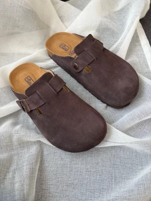 Bruna Birkenstock Boston tofflor mocka - Säljer ett par mörkbruna Birkenstock Boston tofflor i mocka med justerbart spänne och präglad logga på sidan. De har en klassisk täckt tå och bekväm innersula i kork. Perfekta för en chill och stilren vardagslook. Alsrig använda( storlek passar till 37 och 37 1/2) priset kan alltid diskuteras 💖