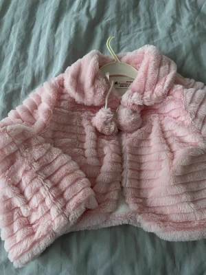 Ljusrosa fluffig pälsjacka med bollar - Supersöt ljusrosa pälsjacka i fuskpäls från Fashion Kids. Jackan har en kort, boxig passform, bred krage och dekorativa bollar i snören framtill. Materialet är mjukt och fluffigt med horisontella ränder för extra struktur. Perfekt för dig som vill ha en gullig och trendig look.
