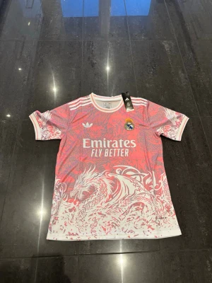 Real Madrid x Adidas rosa matchtröja - Säljer en limited edition Real Madrid matchtröja från Adidas i rosa med vitt drakmönster och klubbmärke på bröstet. Tröjan har vita detaljer och tre ränder på axlarna. Perfekt för fotboll eller träning. Har i alla storlekar