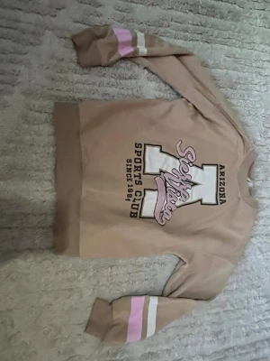 Beige sweatshirt Arizona Sports Club - Beige sweatshirt med tryck 'Arizona Sports Club' och stora bokstäver framtill. Rosa och vita ränder på ärmarna ger en sportig vibe. Mjuk och skön tröja med rund hals och långa ärmar, perfekt för en avslappnad stil.