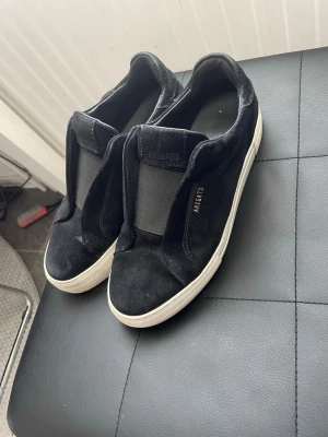 Svarta sneakers från Axel Arigato - Stilrena svarta sneakers från Axel Arigato i mocka med vit sula och diskret guldfärgad logga på sidan. Slip-on modell utan snörning, perfekt för en clean och modern look. Tidlös design som passar till det mesta.