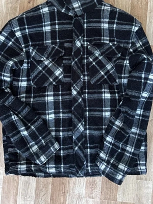 Svartvit rutmönstrad overshirt Bluewear - Overshirt från Bluewear i storlek Large med svartvitt rutmönster. Jackan har två bröstfickor med lock och knäppning framtill. Materialet är mjukt och fleeceliknande, perfekt för lager-på-lager. Snygg och avslappnad stil med klassisk krage.