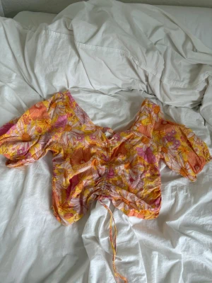 Blommig blus med snörning - Supersöt blommig blus i orange, rosa och gul med kort puffärm och snörning framtill. Croppad modell som man kan anpassa beroende på hur croppad man vill ha den med v-ringning. Perfekt material som är luftigt. 