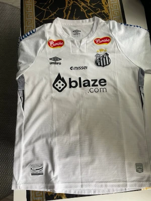 Vit Santos FC matchtröja från Umbro - Snygg vit matchtröja från Santos FC med flera sponsorer tryckta på framsidan, bland annat blaze.com och Canção. Tröjan har klubbmärke på bröstet, rund hals och blå detaljer på axlarna. Tillverkad i lätt och ventilerande material, perfekt för fotboll eller träning.