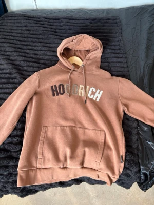 Brun HOODRICH hoodie med huva - Säljer en brun hoodie från HOODRICH med stor magficka och broderad logga i olika färger på bröstet. Hoodien har justerbar huva med snören och ribbade muddar vid ärmslut och nederkant. Perfekt för en chill och avslappnad stil.