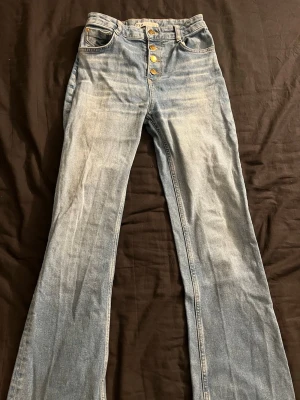 Ljusblå utsvängda jeans med knappar - Snygga ljusblå jeans med utsvängda. Jeansen har fyra synliga guldknappar framtill och klassiska fickor både fram och bak. 