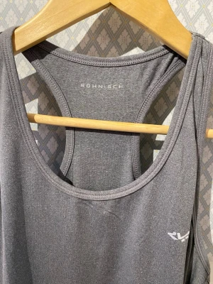 Grå linnetopp från Röhnisch - Snygg grå linnetopp från Röhnisch med sportig design och brottarrygg. Perfekt för träning och aktiva dagar, tillverkad i mjukt och lätt syntetmaterial som andas. Diskret logga på bröstet och enkel passform som ger rörelsefrihet.
