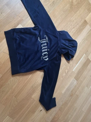 Mörkblå Juicy Couture hoodie - Snygg mörkblå hoodie från Juicy Couture i mjuk velour med huva och dragkedja. Stora Juicy-bokstäver i glittrande stenar på ryggen och diskret logga på bröstet. Perfekt för en chill och trendig look.