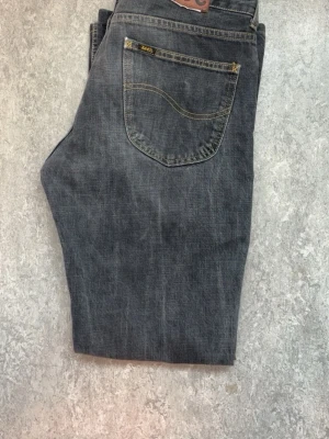 Grå jeans från Lee med gul söm - Säljer ett par grå jeans från Lee med klassisk femficksdesign och snygga gula sömmar. Jeansen har en rak passform och är tillverkade i slitstarkt denim-material. Perfekta för dig som gillar en tidlös och cool look.