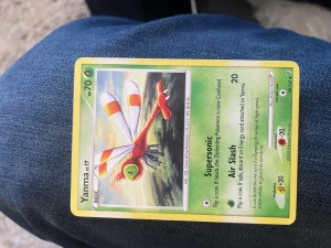 Pokémonkort Yanma 17/147 - Säljer ett Pokémonkort med Yanma, nummer 17/147. Kortet har en grön ram och motivet visar Yanma, en röd och grön trollslända med vita vingar och orange detaljer. Baksidan har klassisk blå Pokémon-design med Pokéboll.