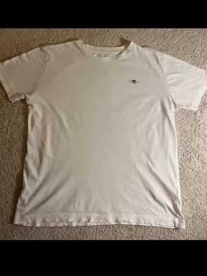 Vit t-shirt från Gant med logga - Klassisk vit t-shirt från Gant med liten broderad logga på bröstet. T-shirten har rund hals och korta ärmar, perfekt basic-plagg i garderoben. Tillverkad i mjuk bomull för skön känsla. Storlek 164, skulle säga som storlek S