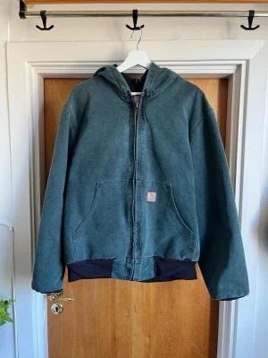Grön Carhartt jacka med huva - Grön jacka från Carhartt med huva, dragkedja och stora framfickor. Jackan har svart mudd vid ärmslut och nederkant samt klassisk Carhartt-logga på fickan. Tillverkad i slitstark bomullscanvas, perfekt för en avslappnad streetwear-look.