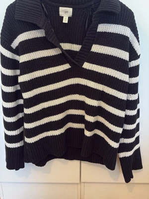 Svartvit randig stickad tröja H&M - Stickad tröja från H&M i svart med vita horisontella ränder. Tröjan har en djup v-ringning, bred krage och långa ärmar. Ribbstickad struktur ger en chunky känsla. Perfekt för en avslappnad och trendig look.