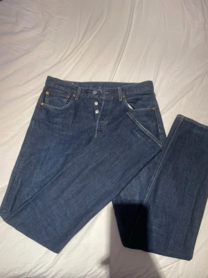 Mörkblå raka Levi's jeans - Säljer ett par klassiska mörkblå Levi's jeans med raka ben och knappgylf. Jeansen har fem fickor, kontrastsömmar och är tillverkade i slitstark denim. Perfekta för en clean och tidlös look.