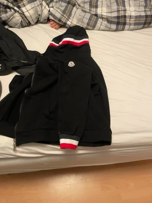 Svart Moncler hoodie med ränder - Svart hoodie från Moncler med dragkedja, huva och snygga röda och vita ränder på huva och ärmslut. Klassisk Moncler-logga på bröstet. Perfekt för en stilren och sportig look.