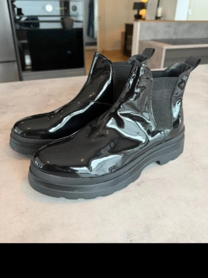 Svarta lackade Chelsea boots - Säljer ett par svarta Chelsea boots i glansigt lackmaterial med chunky sula och elastiska paneler på sidorna. Bootsens höga skaft och dragflikar gör dem enkla att ta på. Perfekta för dig som vill ha en edgy och trendig look.