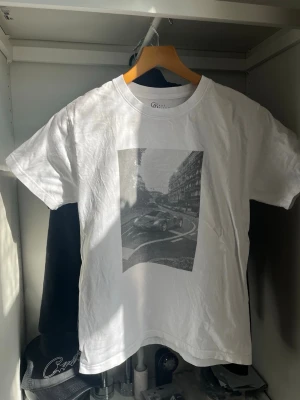 Vit t-shirt från gardegrisch  - Säljer en vit t-shirt med ett svartvitt tryck av en bil och stadsmiljö på bröstet. Klassisk rund hals och korta ärmar. Perfekt för dig som gillar streetwear och snygga prints.