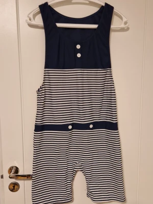 Randig jumpsuit från Panos Emporio i small - Säljer en marinblå och vit randig jumpsuit från Panos Emporio. Modellen är ärmlös med knappar framtill och har ett coolt tryck på ryggen. Tillverkad i mjukt och stretchigt material, perfekt för en avslappnad och sportig stil. Använts en gång vid arbete sen bara legat i garderoben. En knapp återsydd