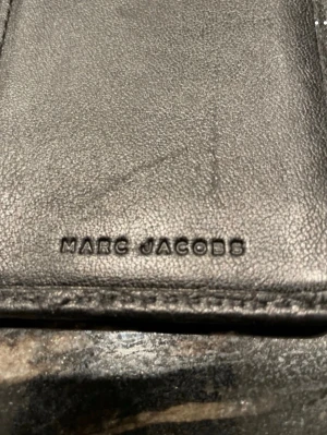 Svart läderplånbok Marc Jacobs - Snygg svart plånbok från Marc Jacobs i äkta läder. Plånboken har flera kortfack och en stilren, minimalistisk design med diskret logga präglad på framsidan. Perfekt för dig som gillar klassiska accessoarer med lyxig känsla.
