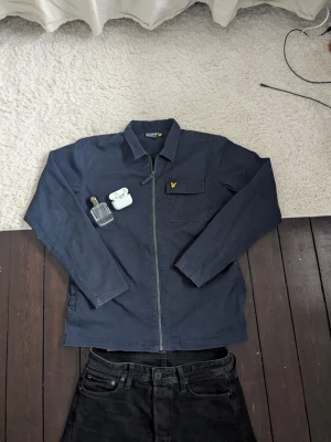 Mörkblå overshirt från Lyle & Scott - Snygg mörkblå overshirt från Lyle & Scott med dragkedja framtill och klassisk krage. Bröstficka med gul logga på vänster sida. y2k. Den är i storlek 14-15 år men är exakt som S