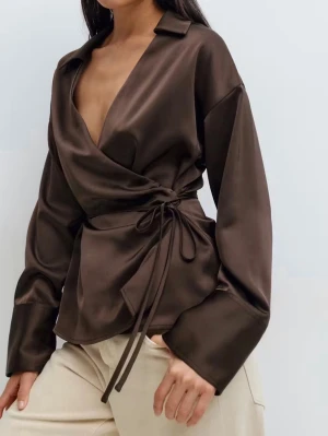 Brun omlottblus i satin - Snygg brun omlottblus i glansigt satinmaterial med knytband i midjan och krage. Blusen har djup v-ringning och långa ärmar med avslappnad passform. Perfekt för en trendig och chic look.