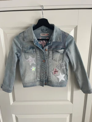 Ljusblå jeansjacka från Desigual - Snygg ljusblå jeansjacka från Desigual med coola stjärnpatches framtill och stort rosa Desigual-brodyr på ryggen. Klassisk krage, två bröstfickor och knappstängning. Perfekt för dig som vill sticka ut med en unik och trendig jacka. PRIS KAN DISKUTERAS VID SNABB AFFÄR!!