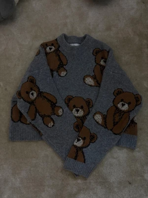 Grå stickad tröja med nallemönster - Superfin grå stickad tröja med stora bruna nallar som motiv. Tröjan har rund halsringning, ribbade muddar och är oversized i modellen. Perfekt för dig som gillar gulliga detaljer och vill ha något mysigt och trendigt i garderoben.