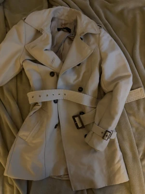 trenchcoat Zara  - Stilren beige trenchcoat från Zara Woman med klassisk dubbelknäppning, bred krage och bälte i midjan. Jackan har detaljerade spännen vid ärmsluten och två sidofickor. Perfekt för dig som vill ha en tidlös och trendig look. Defekt på innertyget men påverkar inte utseende och användning osv.