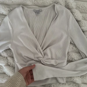 Vit omlottopp med lång ärm - Säljer en vit omlottopp med ribbad struktur och lång ärm. Toppen har en snygg twistdetalj framtill och djup v-ringning. Perfekt att styla med jeans eller kjol för en trendig look.