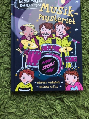 LasseMajas Detektivbyrå: Musikmysteriet - En färgglad och spännande bok ur LasseMajas Detektivbyrå-serien, skriven av Martin Widmark och illustrerad av Helena Willis. Omslaget har tecknade figurer och ett trumset i lila, gult och rosa. Perfekt för dig som gillar mysterier och musik.