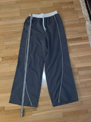Grå baggy träningsbyxor - 34 cm midja, 101 cm längd. Säljer ett par grå tunna mjukisbyxor med vita detaljer och vit resår i midjan samt dragsko. Byxorna har vida ben och är tillverkade i ett material som passar perfekt för chill dagar och gymmet. 