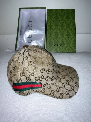Beige GG keps från Gucci - Säljer en beige keps från Gucci med det klassiska GG-monogrammet över hela kepsen. På sidan finns en grön och röd rand som ger den extra Gucci-vibe. Kepsen är i canvasmaterial och har en böjd skärm. Perfekt för dig som vill ha en lyxig och trendig accessoar.