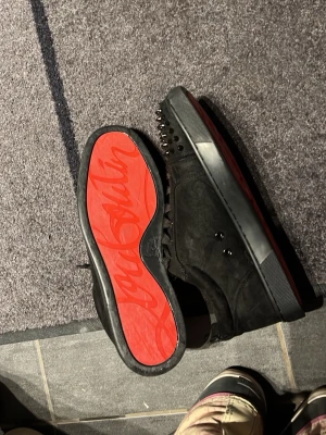 Svarta Cristian Louboutin  - Säljer ett par svarta sneakers från Christian Louboutin med mattsvarta nitar på tån och klassisk röd sula med logga. Skorna är i mocka och har svarta skosnören samt diskreta detaljer längs sidorna. Perfekta för dig som vill sticka ut med en edgy look.