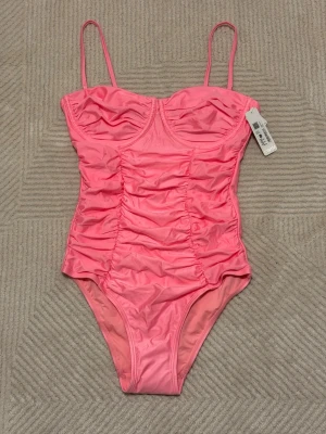 Rosa baddräkt med rynkade detaljer - Trendig rosa baddräkt med tunna axelband och rynkade detaljer framtill. Baddräkten har bygelkupor för extra stöd och är tillverkad i ett glansigt, stretchigt material som ger en snygg passform. Perfekt för dig som vill sticka ut på stranden eller vid poolen.