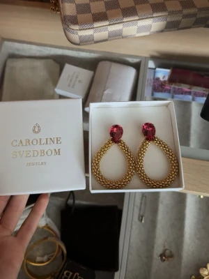 Guldörhängen med rosa stenar från caroline svedbom - Säljer ett par statementörhängen från Caroline Svedbom. De är droppformade i guldton med små pärldetaljer och har stora, gnistrande rosa stenar upptill. Perfekta för dig som vill sticka ut och addera färg till din look. Dem är ej använda utan helt orörda.