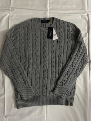 Grå kabelstickad tröja Polo Ralph Lauren - Snygg grå kabelstickad tröja från Polo Ralph Lauren med rund halsringning och ribbade muddar. Tröjan har det klassiska broderade logotypen på bröstet och är tillverkad i mjukt material. Perfekt för en stilren och avslappnad look.